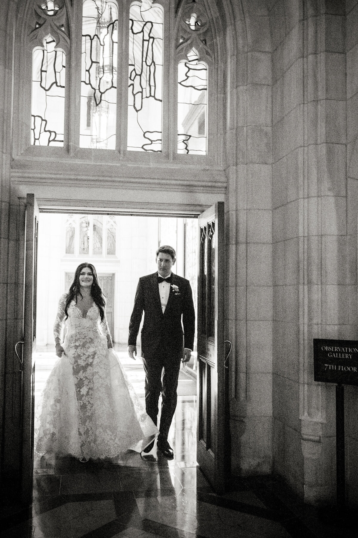 The stunning photo captures a moment at Washington D.C.'s iconic Washington National Cathedral, beautifully shot by photographer Twah Dougherty, with Current Films as videographers. In the image, a bride dressed in a lovely lace wedding gown and a groom in a classic tuxedo are seen walking hand in hand through a grand arched doorway. Above them, intricate stained glass windows adorn the scene, adding a touch of elegance. The couple is entering a room with breathtaking Gothic architecture, and a visible sign reads Observation Gallery 77th Floor, hinting at the breathtaking view that awaits them.