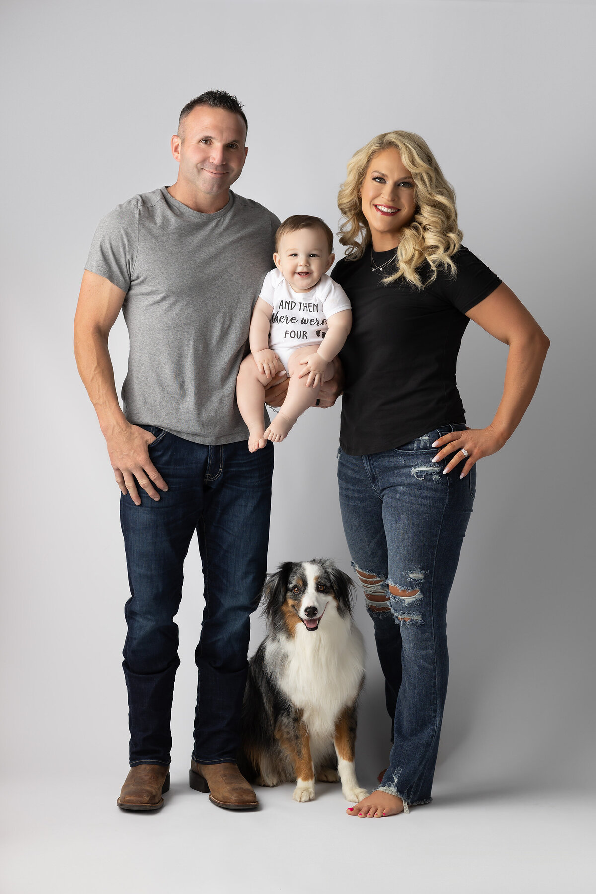 DFW milestone photography of a smiling family of four—mom, dad, baby in a "And Then There Were Four" onesie, and their Australian shepherd—posed against a soft gray backdrop.