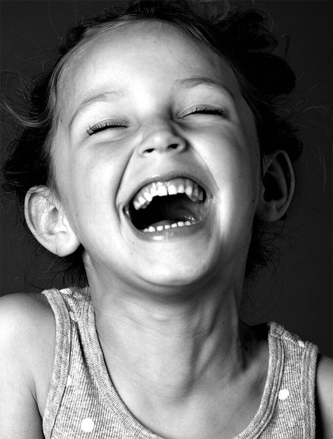 A close-up black and white photo captures a young girl wearing a polka-dot tank top, eyes closed and mouth wide open in joyful laughter, exuding pure happiness.