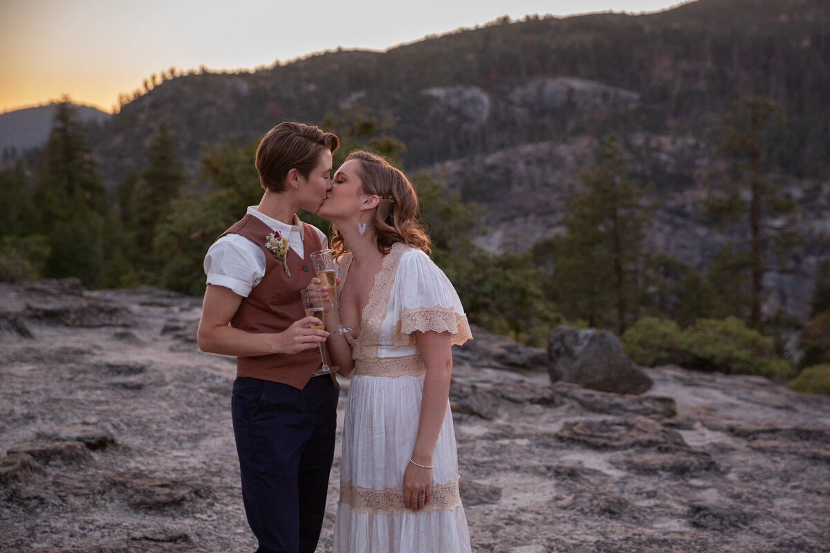 Two brides kiss while holding champagne glasses on their elopement day.