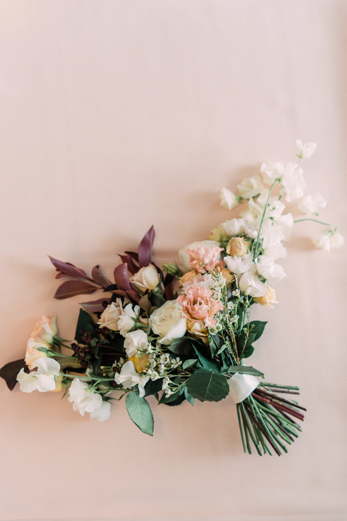 Bouquet of red foliage with white sweet peas & roses, waxflower, & pink lisianthus on pale pink background at Cornman Farms