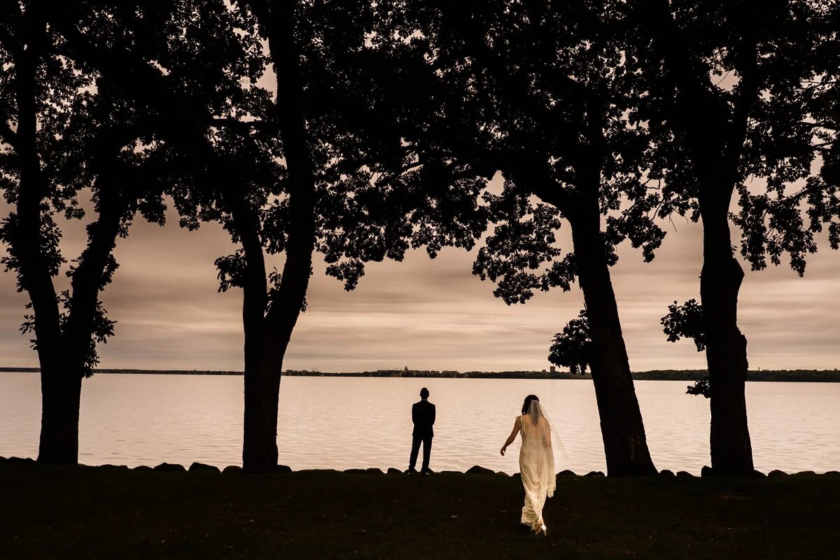 An ethereal first look scene by a lakeside, with the bride approaching the groom under a canopy of trees in silhouette.