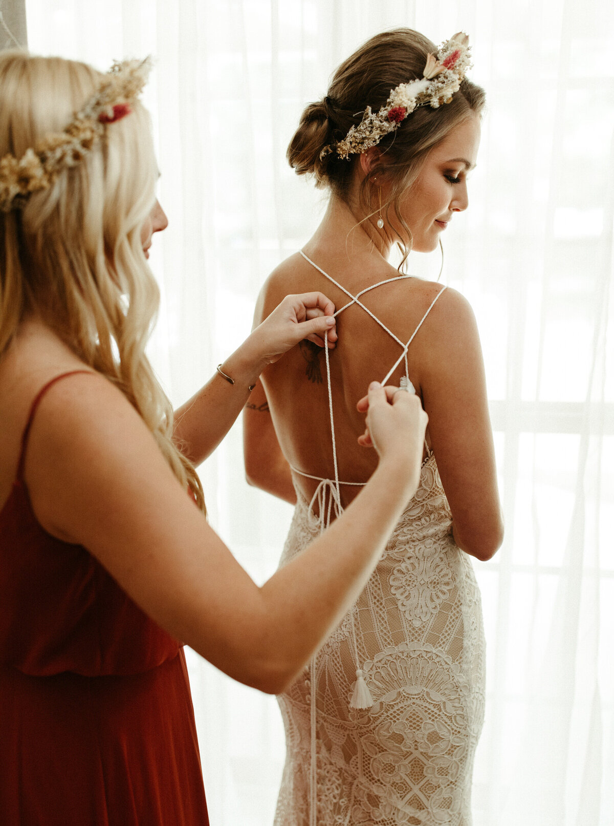 Maid of honor in flower crown helping tie the intricate straps on bride's wedding dress