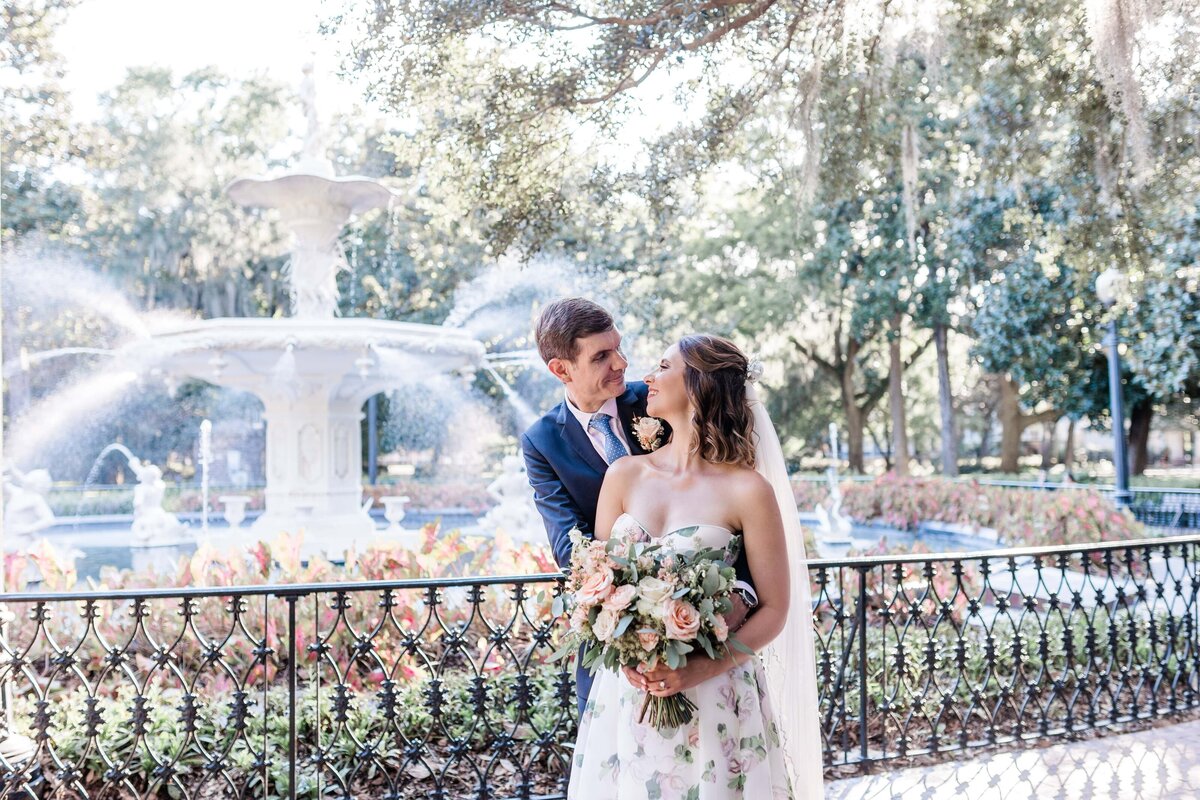Groom embracing bride holding a pastel bouquet, with the Forsyth Park fountain in Savannah, GA, as the backdrop during their elopement.