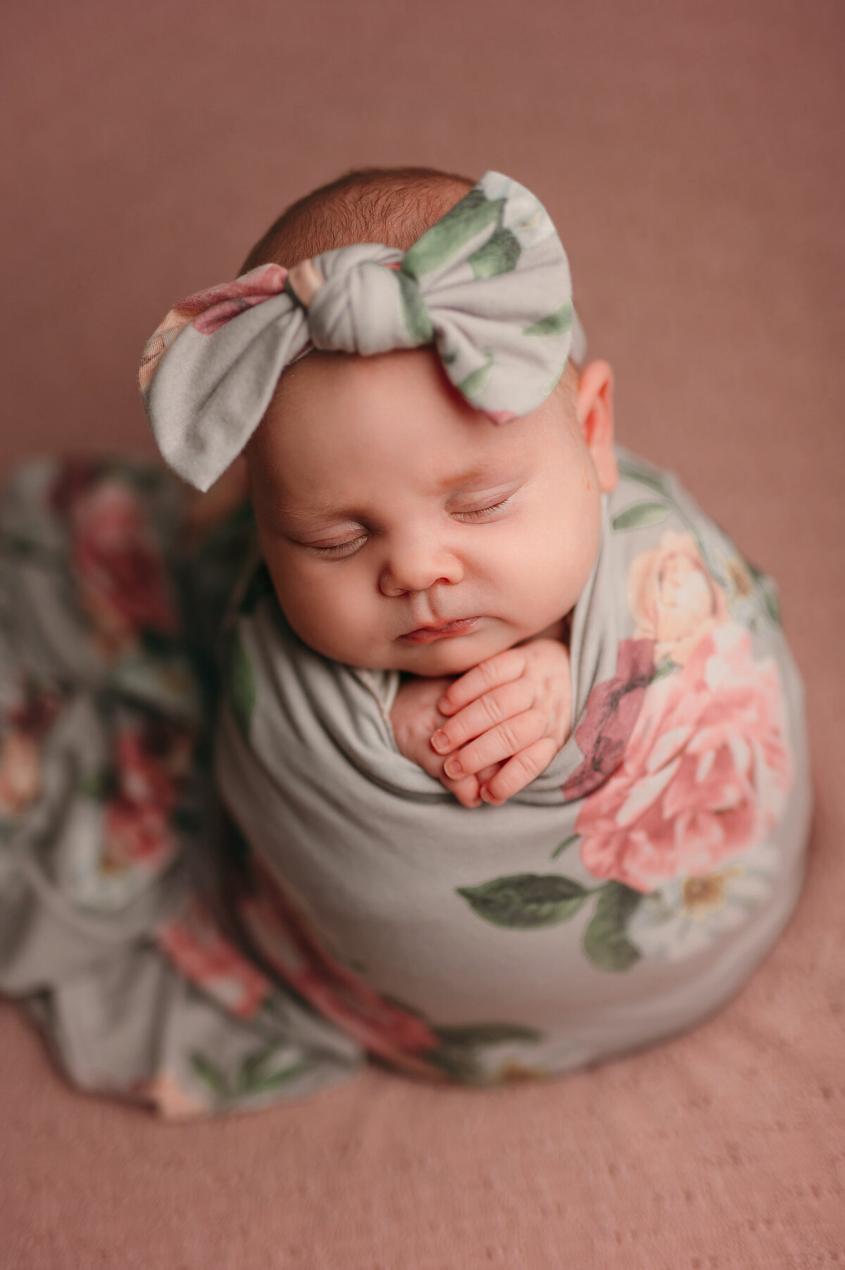 Portrait of baby girl swaddled  like a cocoon in a floral wrap with matching headband against a pink backdrop.
