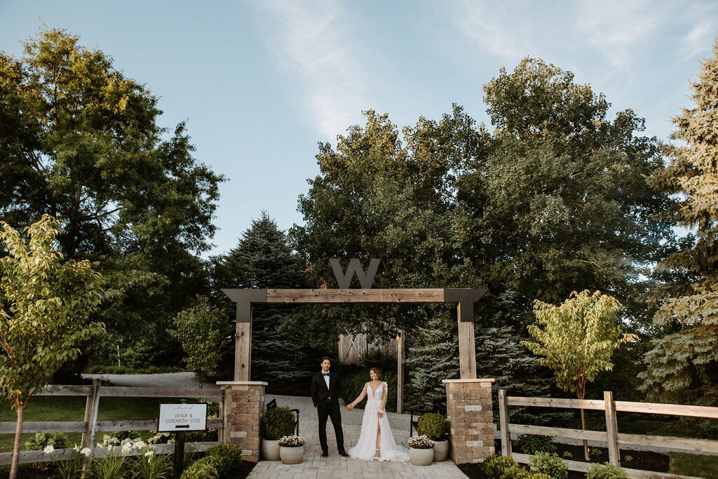 bride and groom standing under the entrance arch of Willowbrook wedding venue with lush landscaping and trees