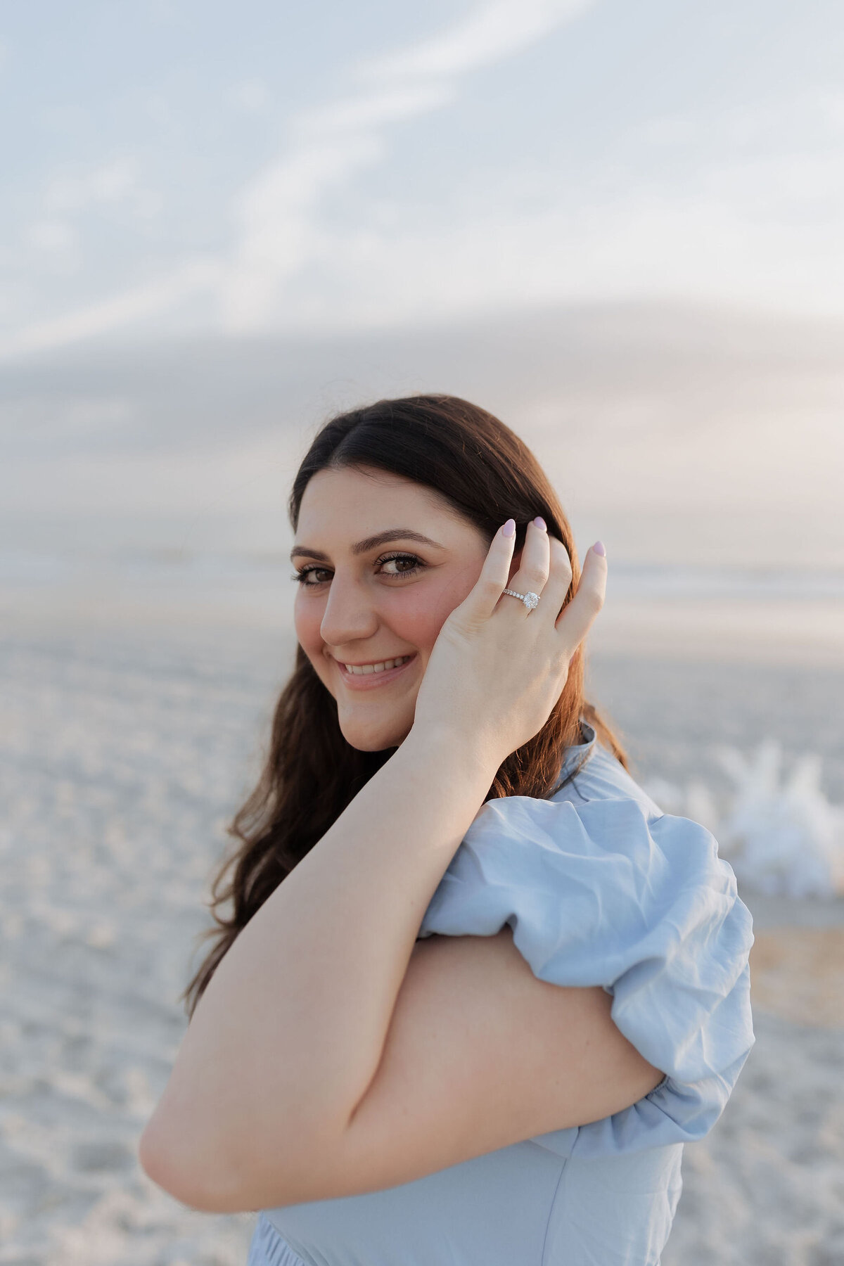A bride-to-be smiles sweetly at the camera while gently tucking her hair behind her ear to reveal her engagement ring during a beach session at sunset. Dressed in a soft blue outfit, she stands against a peaceful shoreline with pastel skies in the background. This joyful and radiant moment was captured by Heartful Films, a Jacksonville engagement photographer and videographer known for elegant, true-to-color portraits that celebrate love naturally.