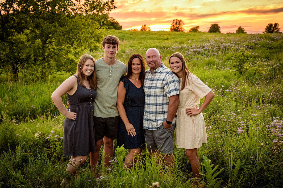 Family portrait with three adult children and their parents standing in a long grassy field with sunset in background in Fonferek Glen County Park near Green Bay, Wisconsin