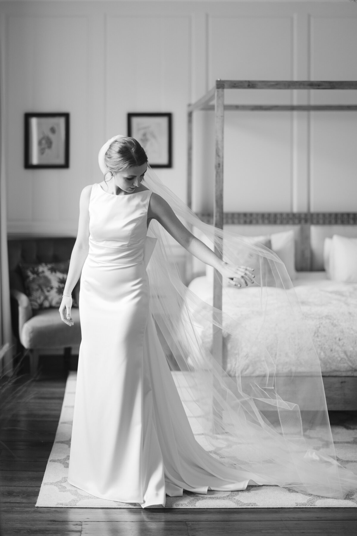 A bride in a sleek, elegant gown and long veil stands in a softly lit Syrencot Wedding suite with a four-poster bed. She gazes down, gently holding her veil. The room features framed artwork and a plush chair, creating a classic, serene atmosphere. Black and white image.