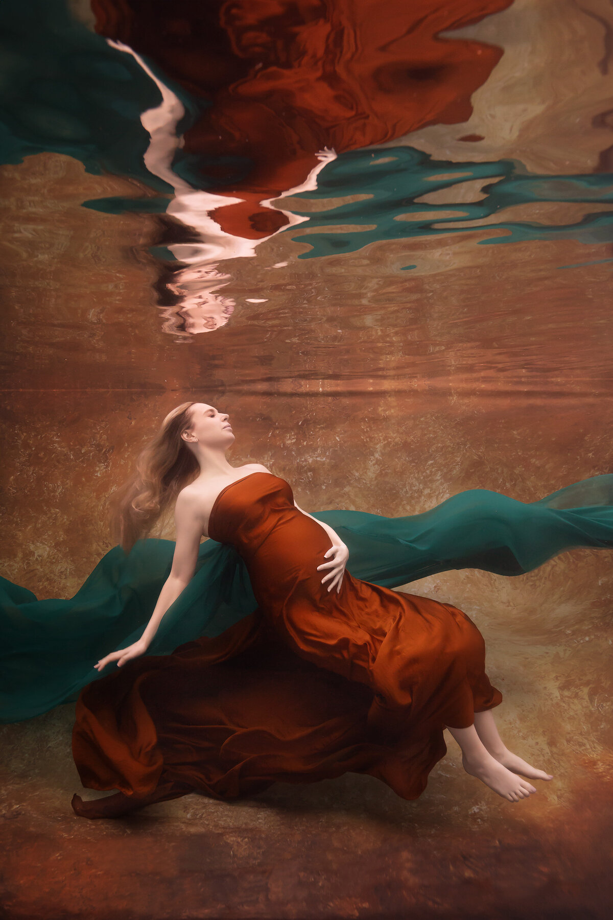 A beautiful underwater maternity photo captured by Dallas underwater maternity photographer Lindsay Walden. The woman floats gracefully in a rust-colored gown with flowing teal silk drifting around her, creating a harmonious contrast against the water's warm tones. Her serene expression adds a dreamlike quality to this underwater maternity photoshoot.