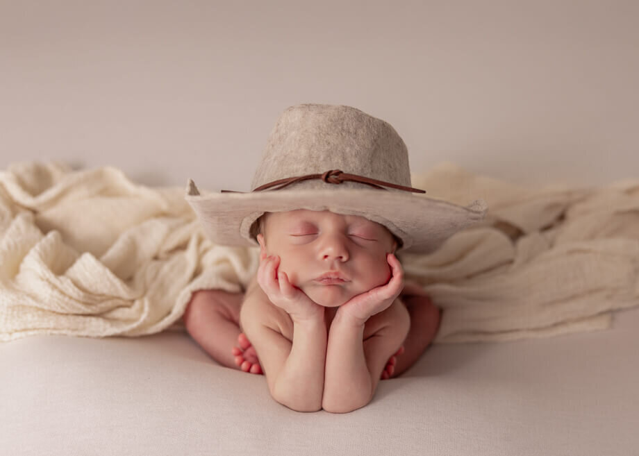 froggy pose with cream background.  Baby boy is wearing a cowboy hat and has a drape over his back