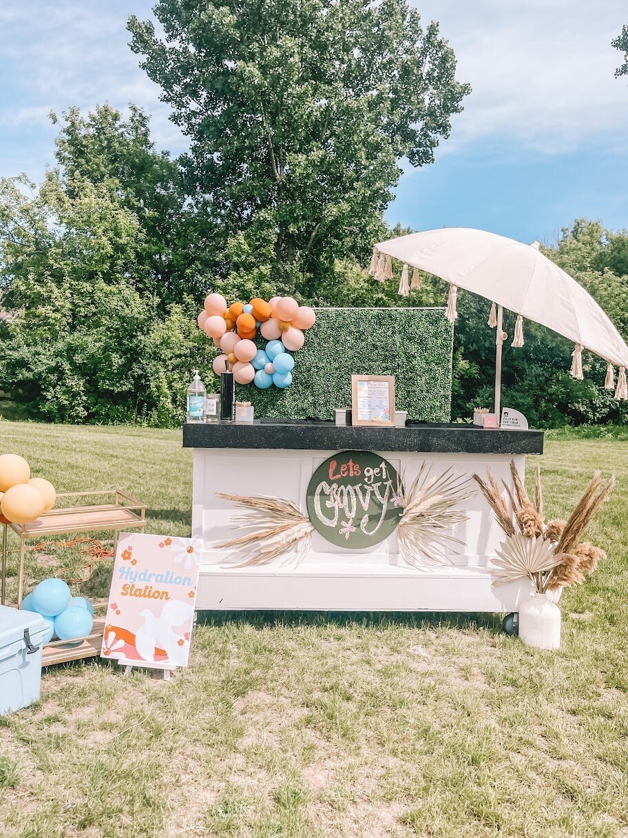 A mobile bar cart set up outside with an umbrella, colorful balloons, and pampas grass.