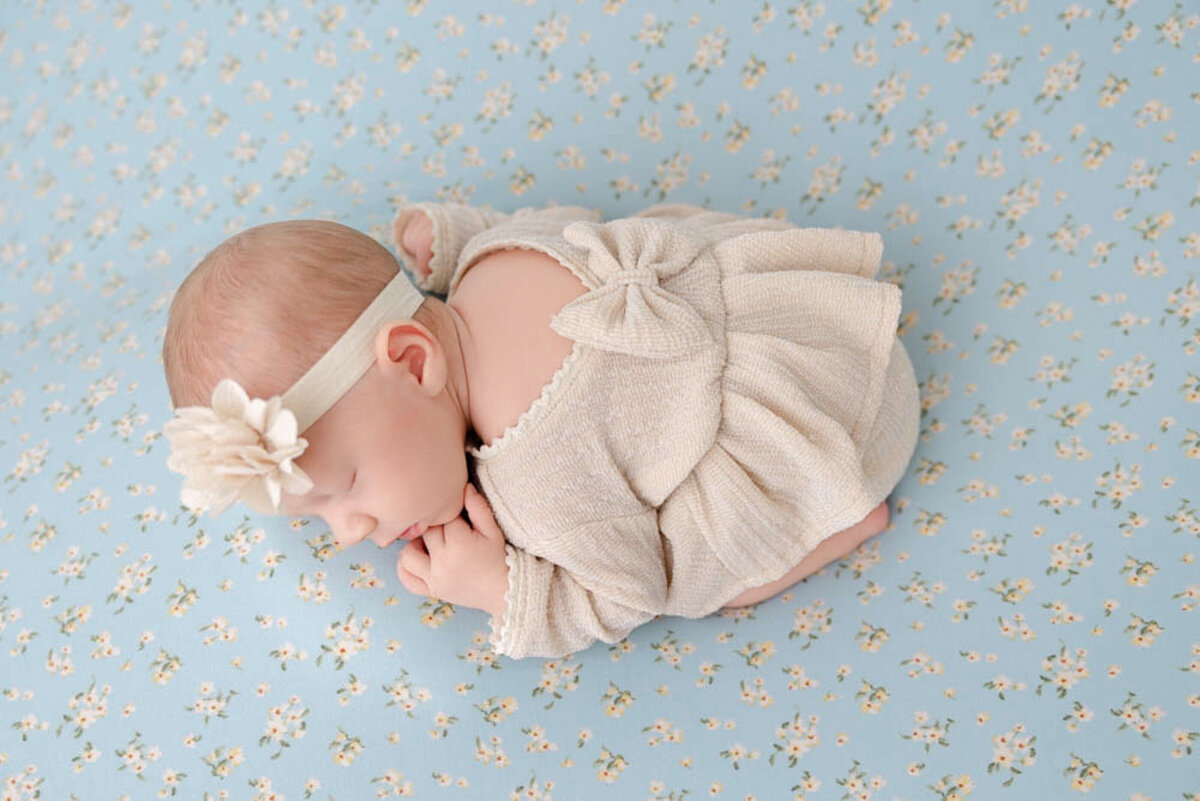 Baby girl newborn posed on a blue flowered blanket and wearing a champagne colored dress with a bow on her back and a cream-colored headband.
