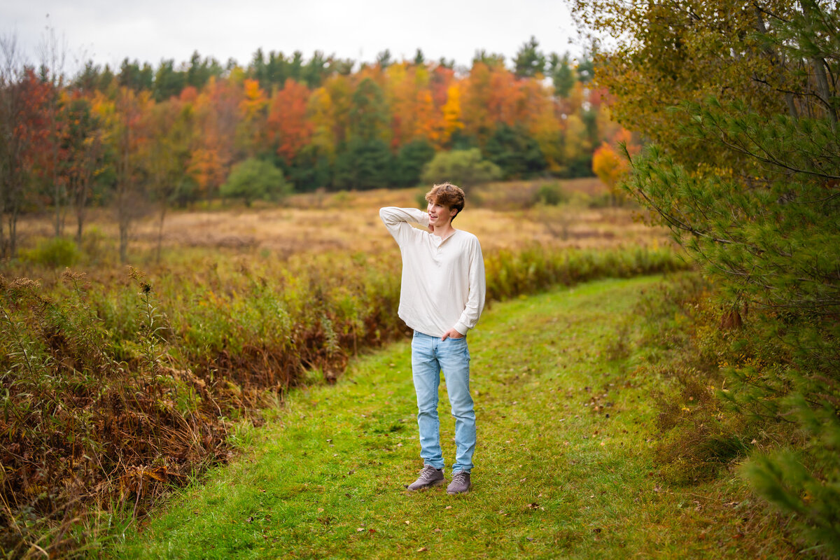 High school senior boy standing in a grassy field with autumn trees.