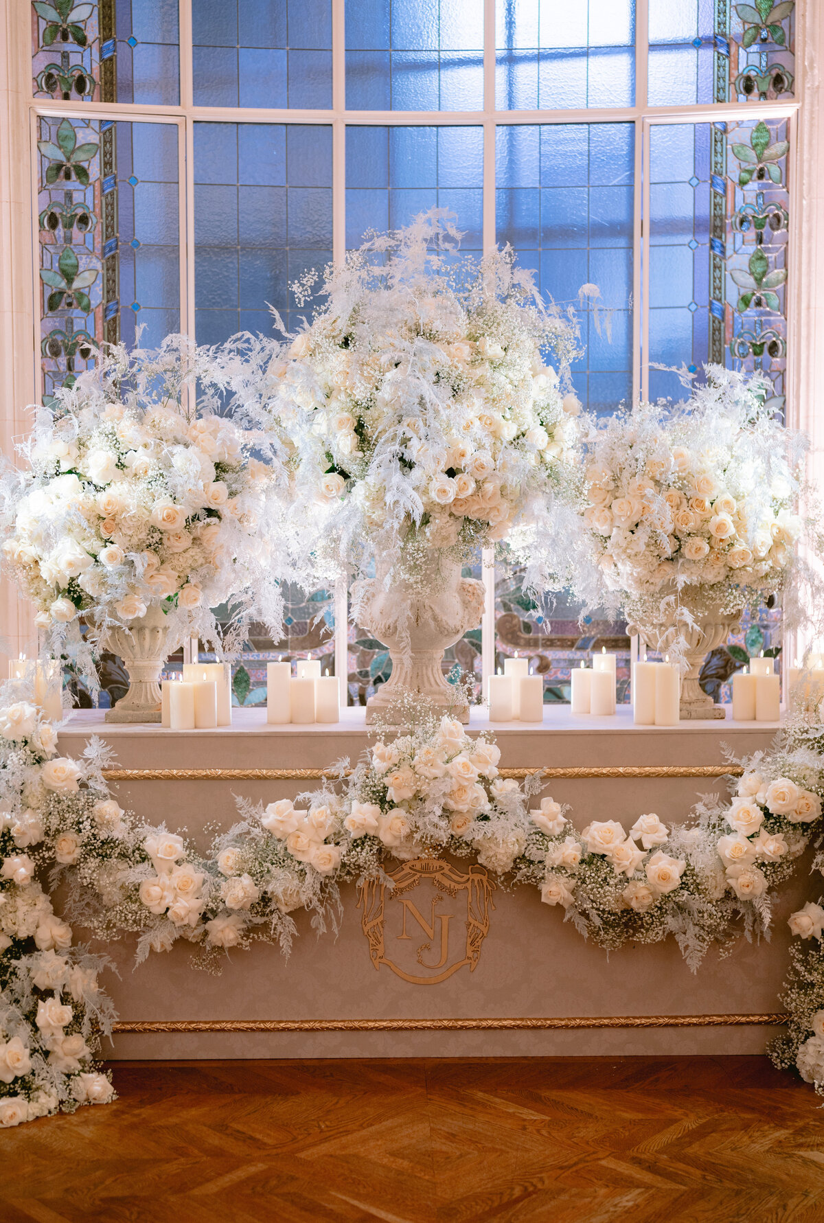 The stunning photo was captured at two luxurious locations, the Shangri La Hotel and Ritz Paris Hotel in Paris, France, adding an extra touch of opulence to the already enchanting scene. Renowned photographer Matteo Coltro skillfully mastered the art of capturing the elegance and beauty of the decor. Zen Film Works also contributed to capturing the enchanting scene, showcasing their expertise as videographers alongside Matteo Coltro's photography.  An elegantly decorated room showcases a grand display of white and ivory roses, ferns, and various floral elements, elevating the aesthetic with a touch of natural beauty. Positioned on a mantle beneath a splendid stained glass window, a cluster of white candles illuminates the space, enveloping it in a soft and romantic glow.