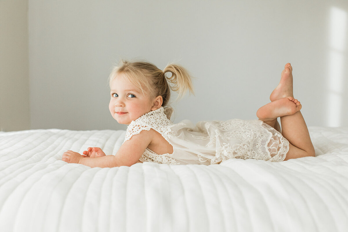two year old girl in white dress lays on bed and looks at camera with a smile
