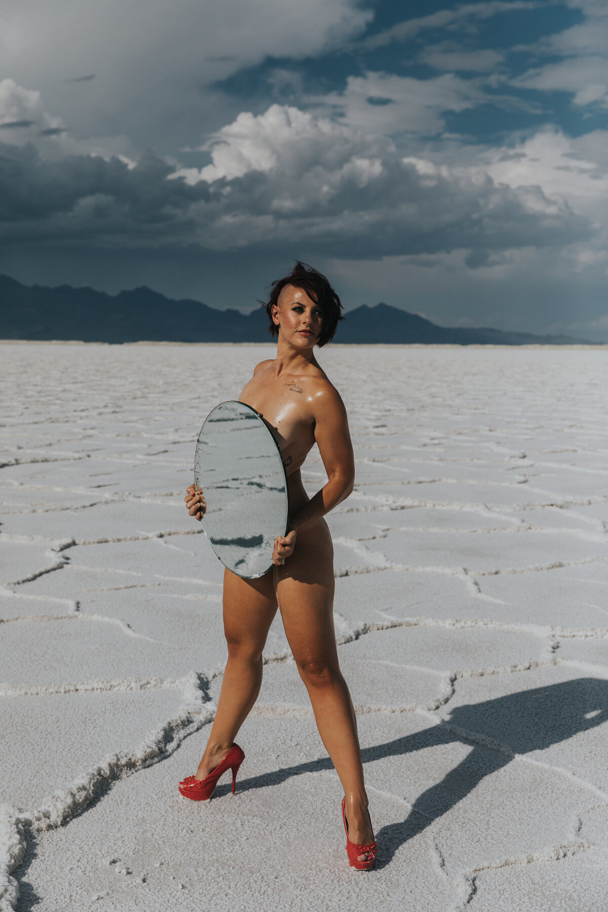 A woman standing in onlyl red heels and holding a mirror infront of her.