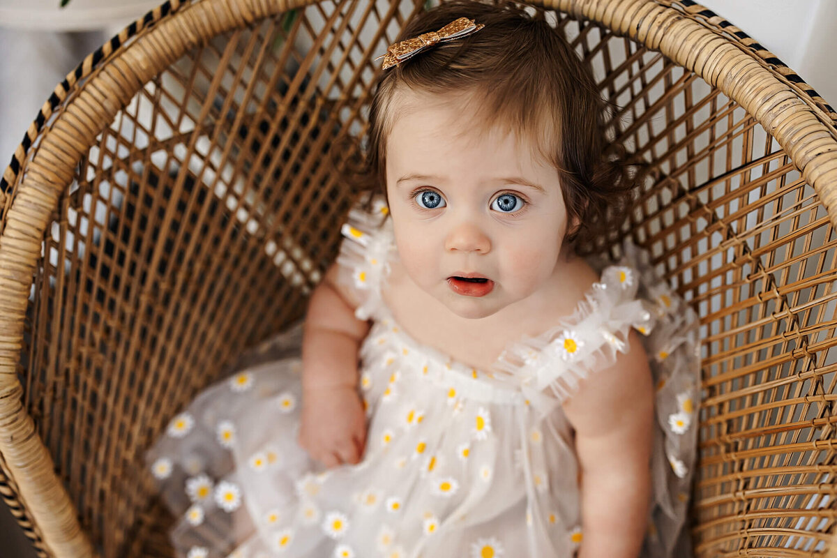 A baby with blue eyes and curly brown hair sits in a wicker chair, wearing a white dress with small yellow daisies and a bow hair clip, looking up with a curious expression—perfect for milestone celebration portraits.