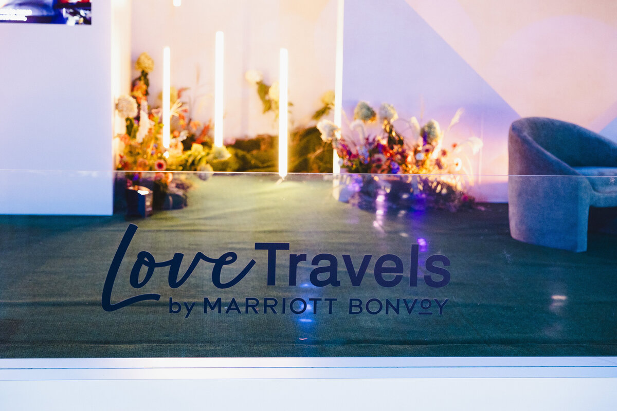 A sign says Love Travels by Marriott Bonvoy with florals and lights behind.