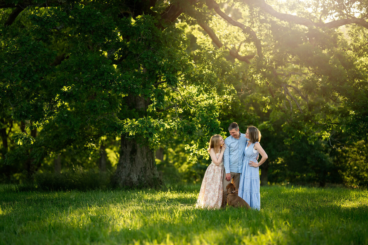 A family of three and their dog stand in the sunlight by a tree on the Biltmore Estate. 