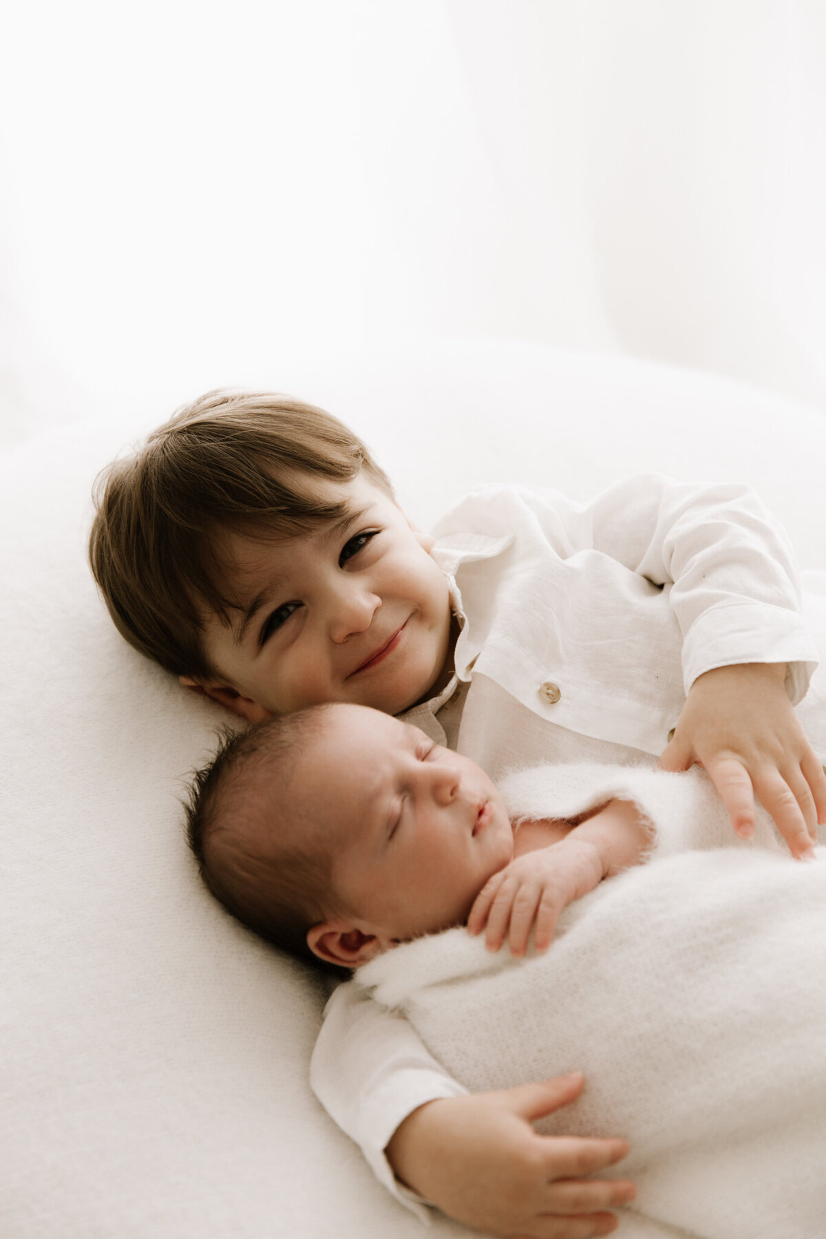 A smiling toddler lies beside their newborn sibling, both wrapped in soft white textures, sharing a peaceful moment on a cream blanket in a softly lit studio.