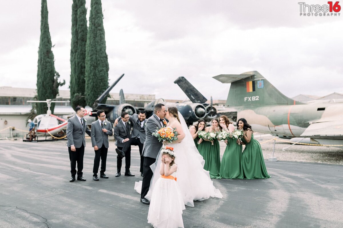 Bride and Groom share a kiss as the Bridal Party acts silly and Flower Girl covers her eyes