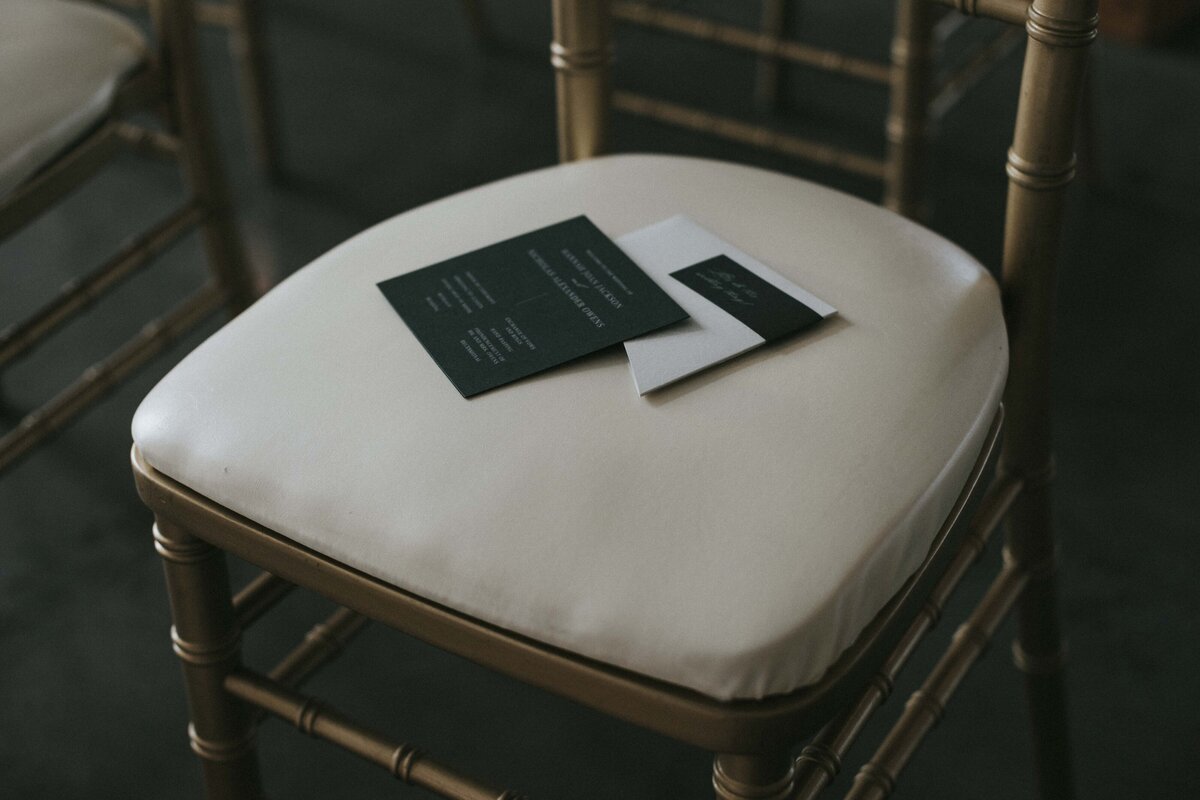 Dark green wedding invitation with white font and white asymmetrical envelope set atop a gold and white elegant chair.