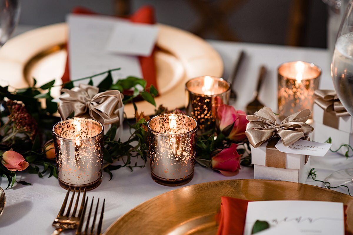 A table set with a white table cloth, gold chargers, gold flatware and gold votive candles. A garland of greenery accented with orange tea roses runs down the center of the table.