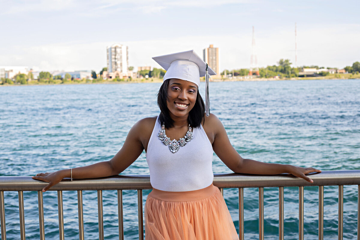 Detroit Riverwalk Senior Photoshoot | Detroit Michigan Senior Photographer, Senior girl with long peach tutu skirt, white sleeveless top, and white graduation cap leaning on rail overlooking the Detroit River