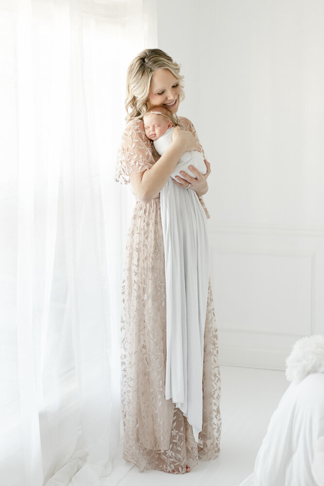A blonde mother snuggles her newborn baby standing in a white room in kristie lloyd's nashville photography studio