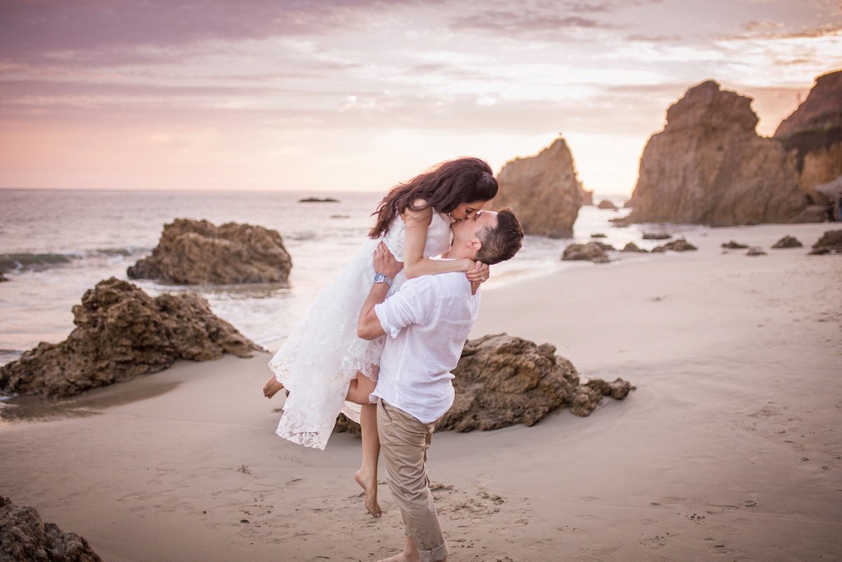 Groom to be lifts his Bride up in the air and kisses her while standing on the beach
