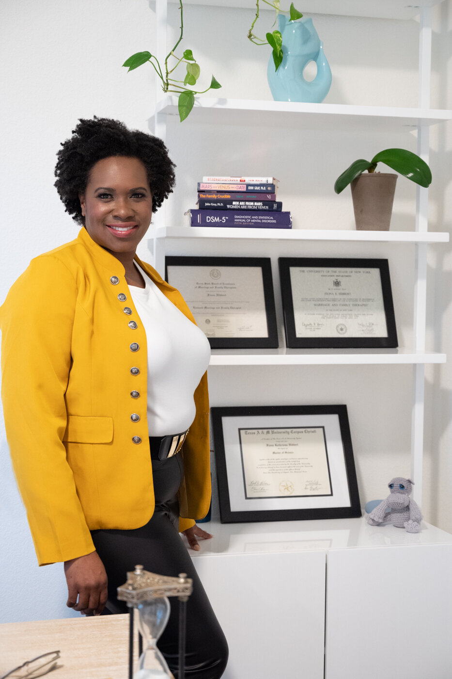 A professional image of a black woman wearing a yellow jacket and black pants standing next to her school certifications smailing to the camera