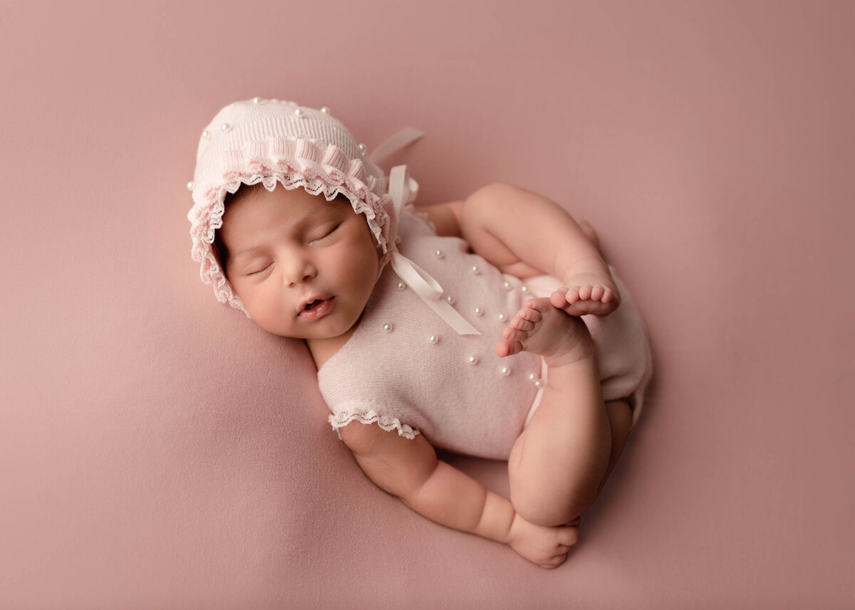 Baby Sleeping posed on back on pink knit blanket. Wearing blush onesie and bonnet with lace and pearl accents.