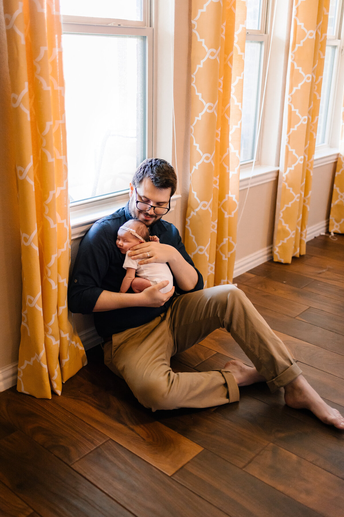 Experience professional maternity portraits in Albuquerque with this serene image of a man holding his newborn baby. Seated on a wooden floor, the man is dressed in brown pants and a dark blue shirt. The background features yellow and white curtains, adding a soft and warm touch to the scene.