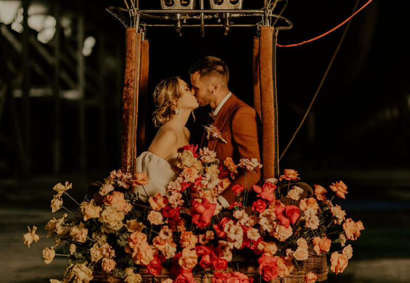 Mariés dans une montgolfière débordant de fleurs aux teintes roses et corail, échangeant un baiser lors d'une séance photo mariage.