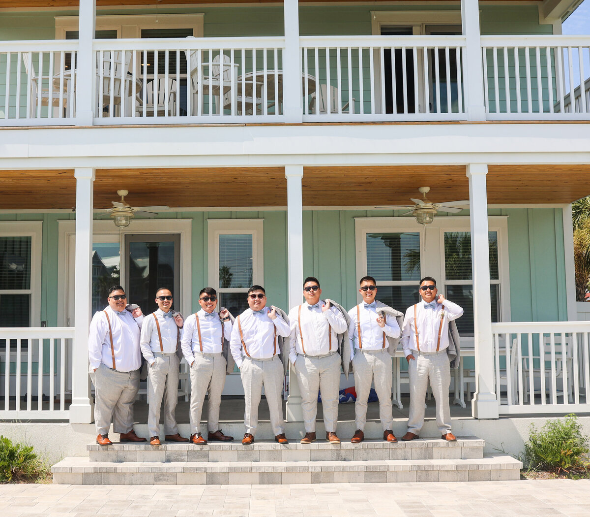 groom and groomsmen wearing brown suspenders and holding suit coats over shoulder and wearing sunglasses in front of blue beach house in Panama City Beach Florida
