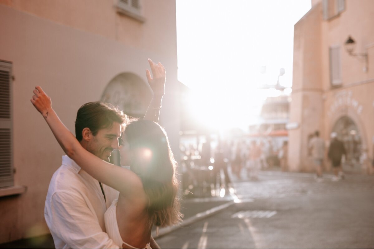 Woman throwing her arms around her fiances neck with the streets of France behind them and the sun about to set.