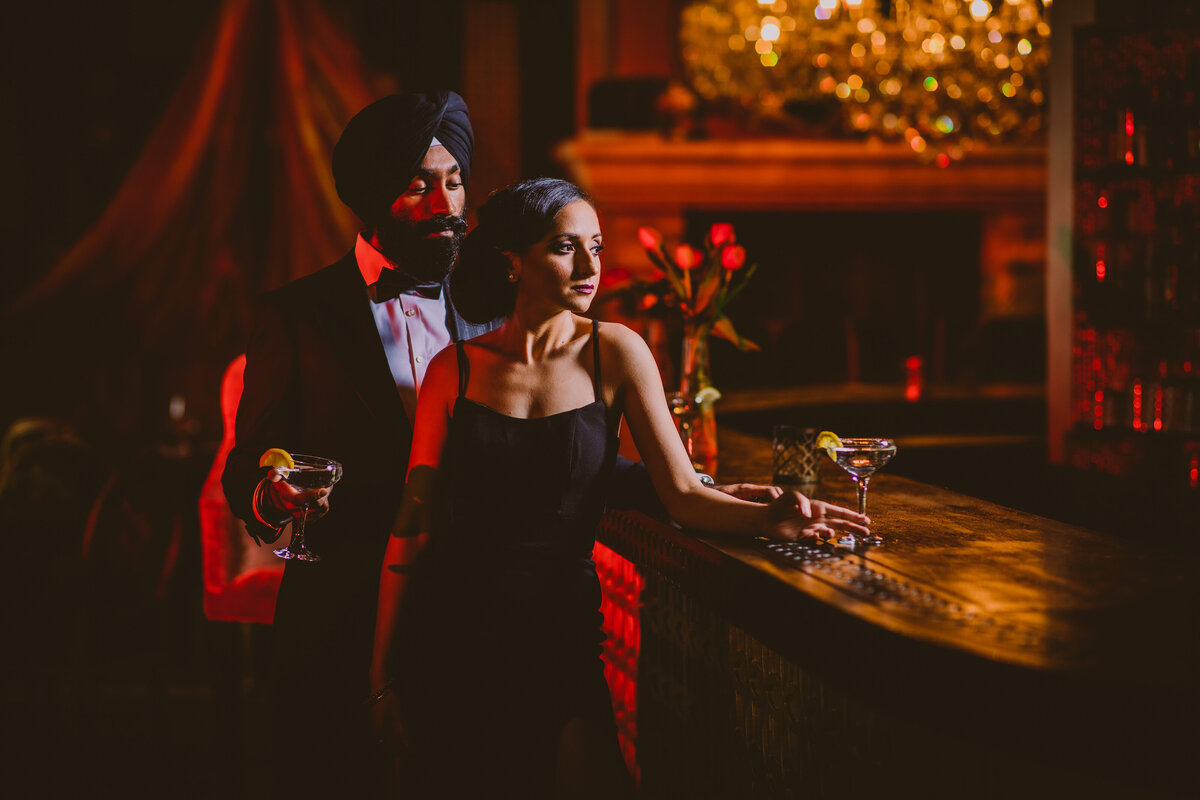 Engaged couple in black formal outfits having drinks at a new york city bar indoors in a dark, red moody setting