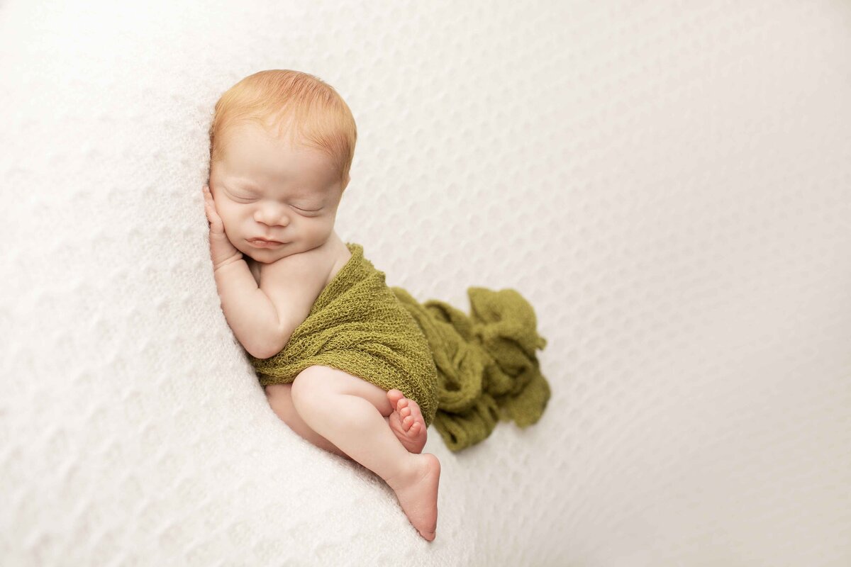 Peaceful newborn baby boy with red hair posed on his tummy, his tiny hands tucked under his cheek on a soft backdrop