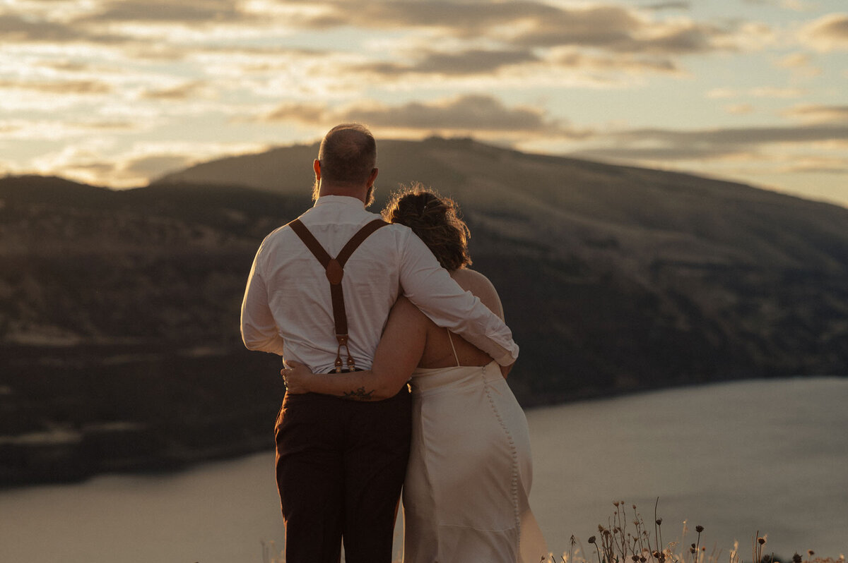bride and groom wearing wedding attire hold each other while looking out at the scenery of the Dalles, Oregon under a sunset sky