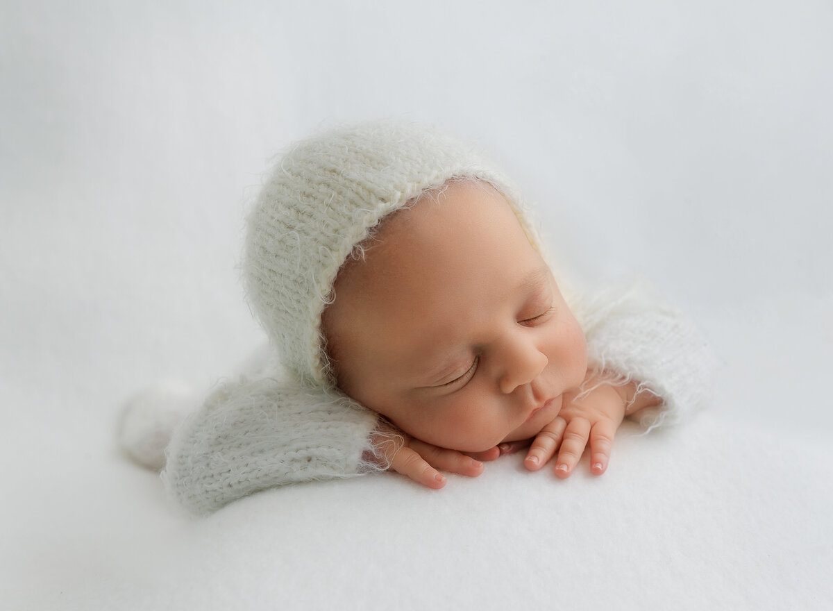 Baby is sleeping in a white knit romper and hat for their brooklyn ny newborn photoshoot. Baby is sleeping on his belly with his hands folded under his chin. Captured by top Brooklyn newborn photographer Chaya Bornstein