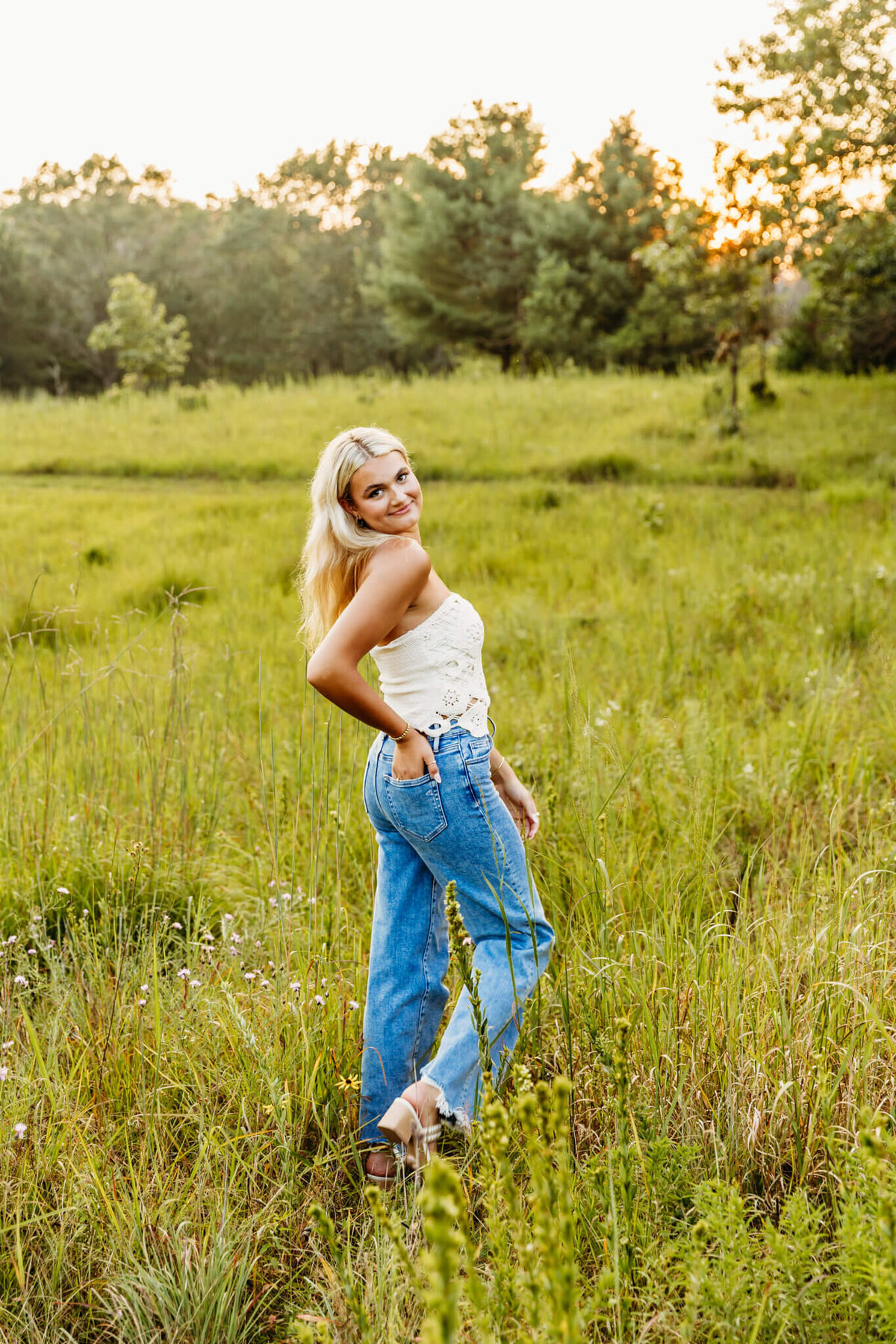 young lady in white top and jeans walking through a grassy field in Neenah and looking back as she puts her hand in her pocket. 