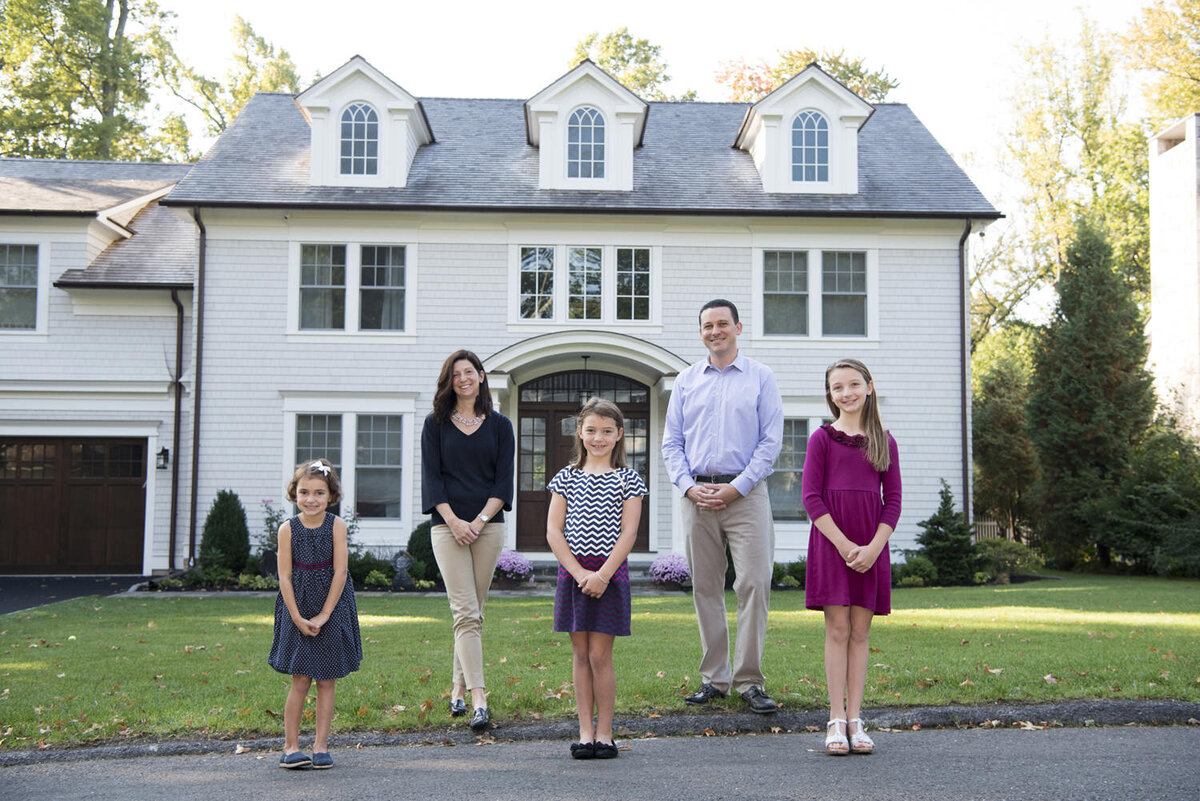 A family of five standing together in front of their house with their arms in front of themselves 