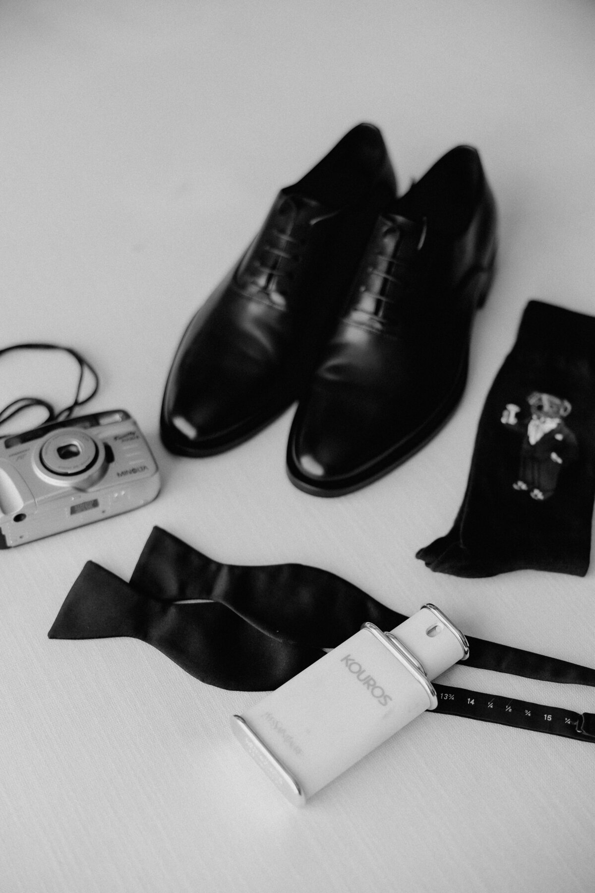 Groom details on a clean white linen couch in hotel room, including shoes, socks, cologne, bowtie and film camera