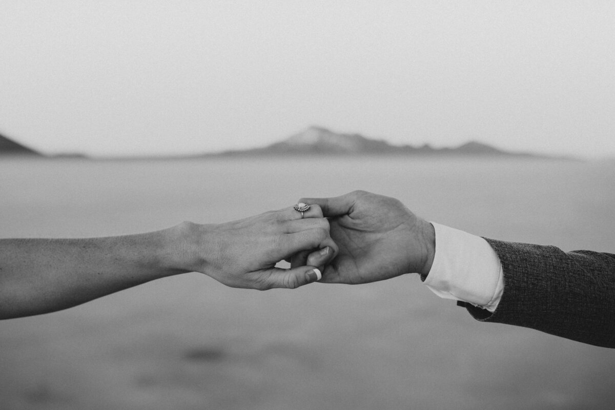 A close-up, black and white photo of a couple's hands tenderly holding each other with mountains in the distance