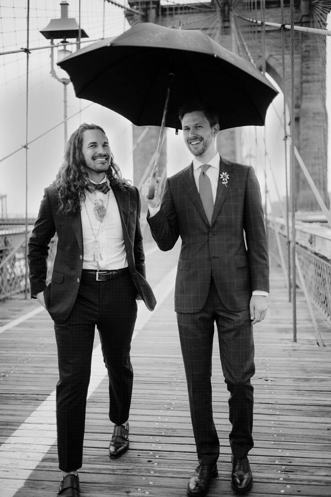 The two grooms are walking on the Brooklyn Bridge, under a black umbrella. NYC City Hall Elopement Image by Jenny Fu Studio