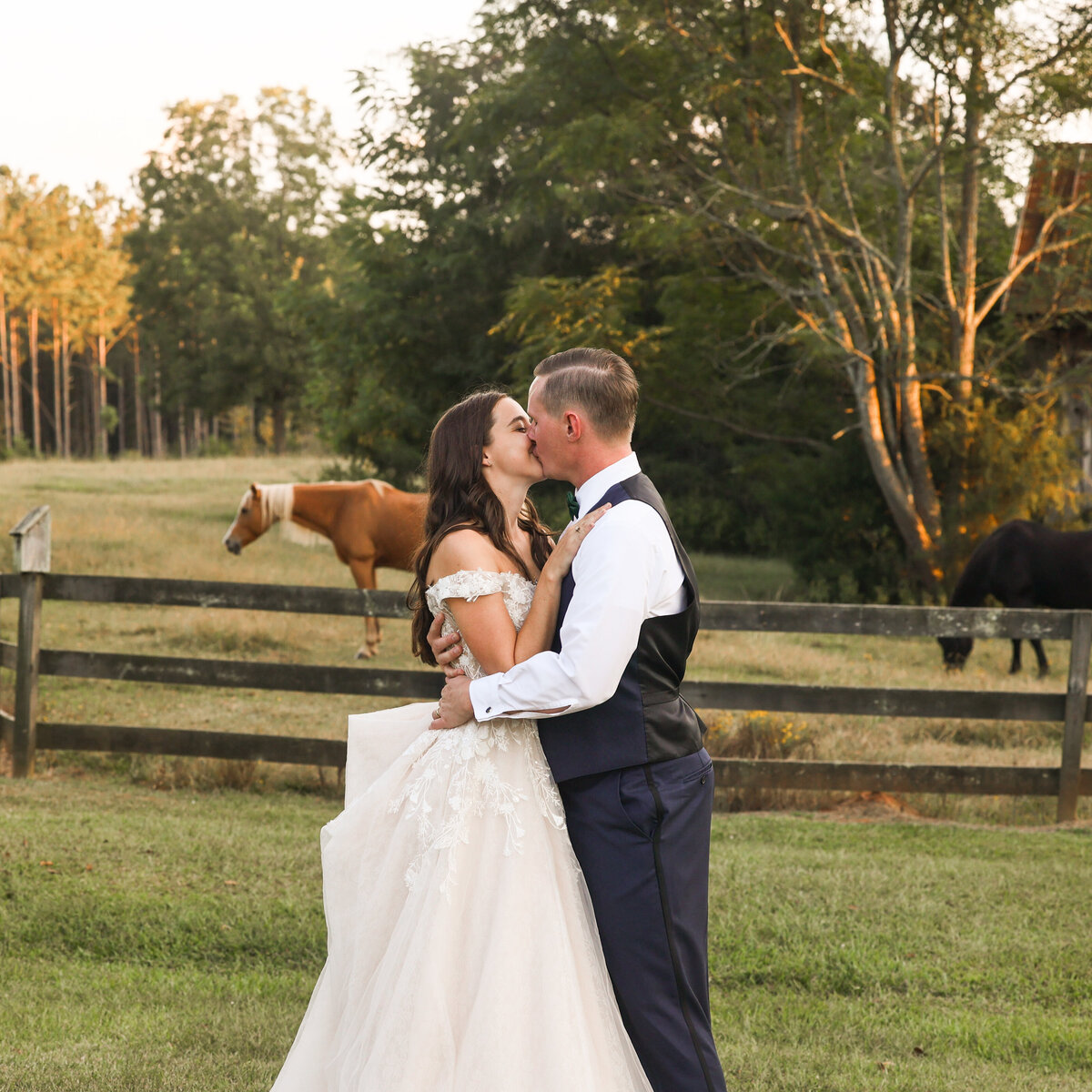 bride wearing white tulle dress and groom wearing blue suit and white shirt kissing with horses in the background