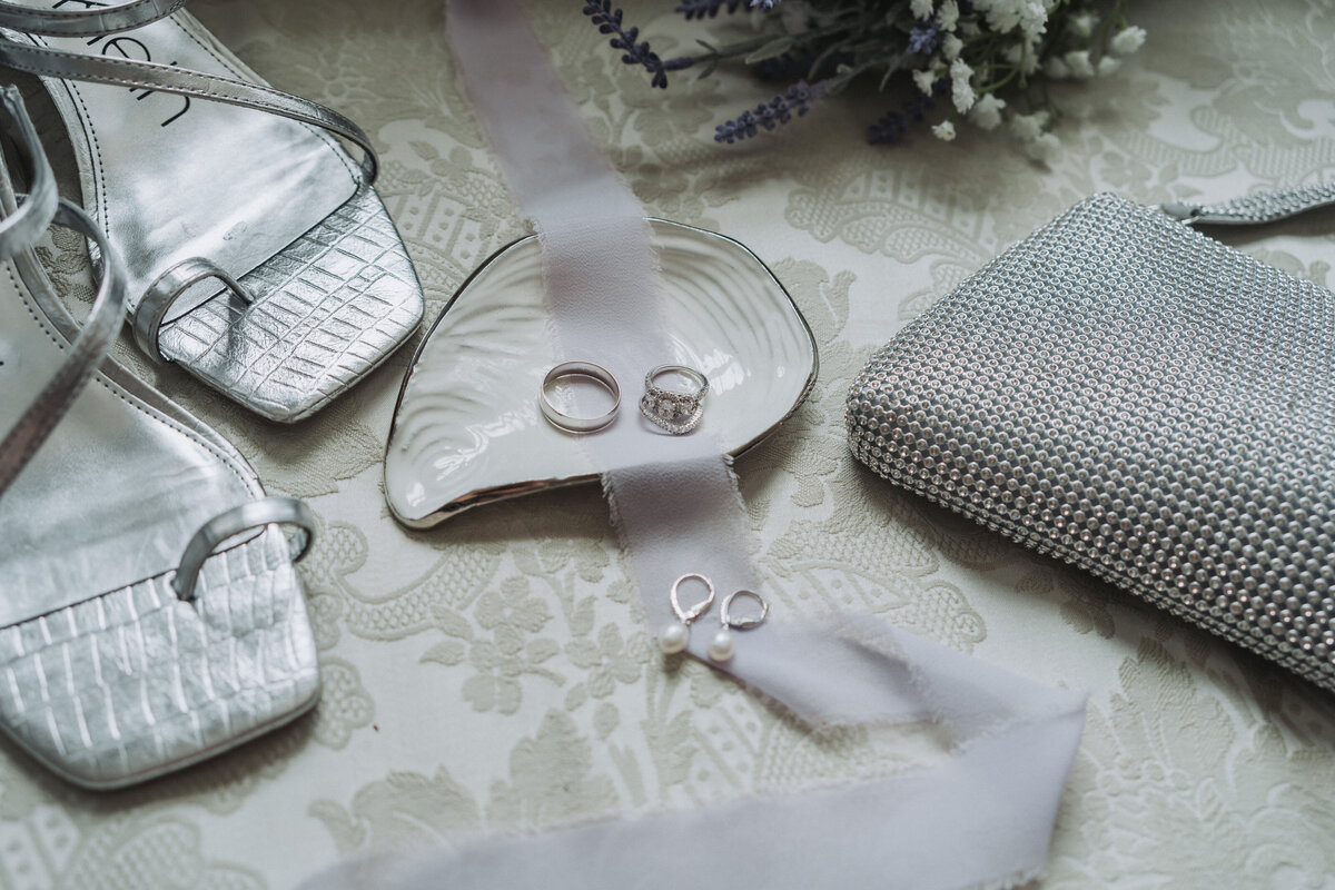 Close-up of sparkling wedding rings on a white shell dish with a lavender ribbon, surrounded by a bridal bouquet and other elegant details at the historic Obici House in Suffolk, Virginia, photographed by Quinn Photography LLC.