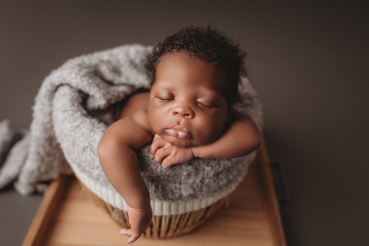 Sleeping newborn boy with dark curly hair propped in a woven basket with his chin on his hand