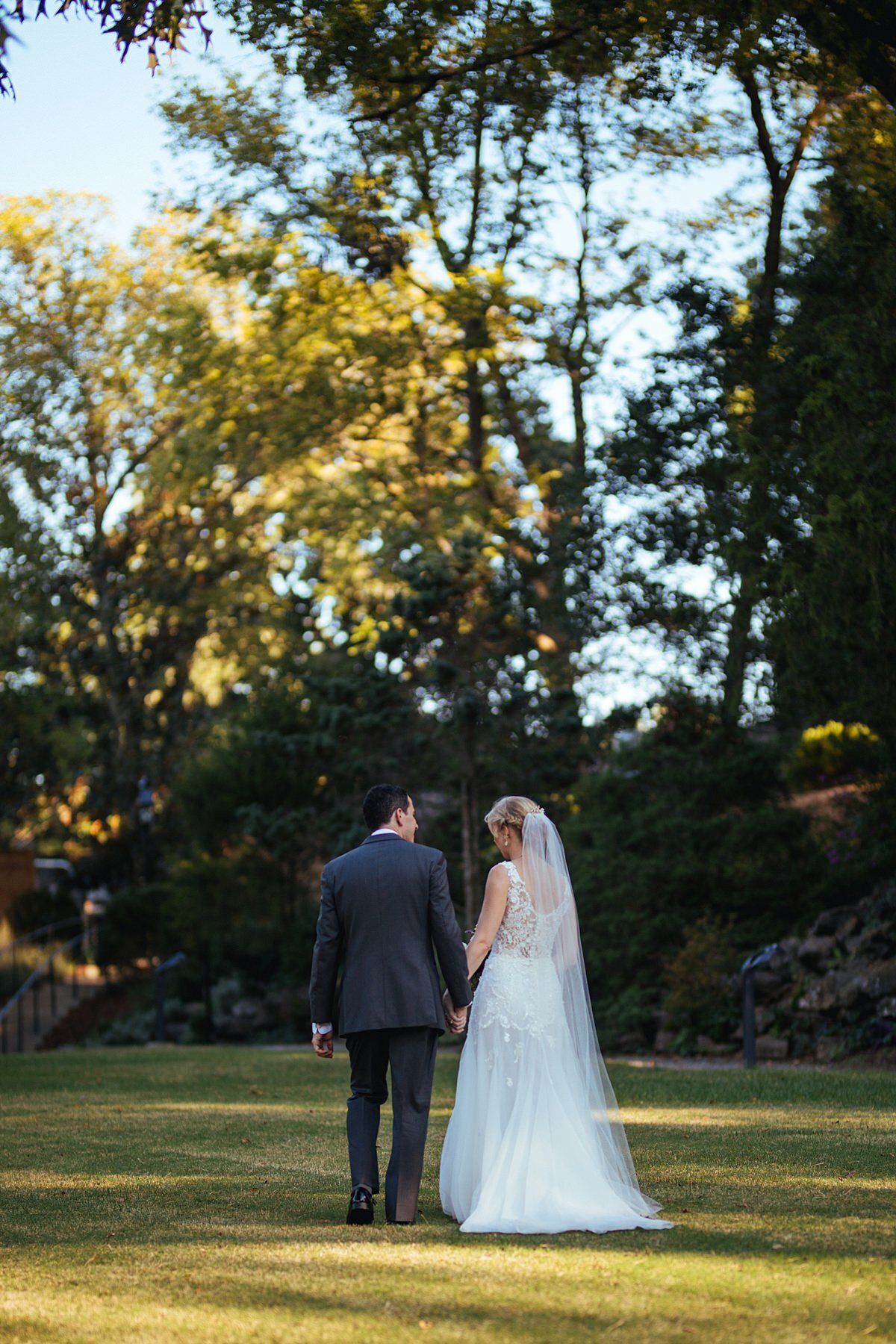 the bride, wearing a long sleeveless lace wedding dress and long sheer veil and the groom wearing a charcoal gray tuxedo hold hands as they walk away through the garden at Cheekwood